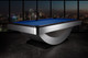 Three-quarter view of the Doc & Holliday Poshy Reverse Rainbow Slate Pool Table with Euro Blue Championship felt, matte black upper rails, and brushed metallic base, staged on a concrete floor with a modern backdrop wall. Three-quarter view of the Doc & Holliday Poshy Reverse Rainbow Slate Pool Table with Euro Blue Championship felt, matte black upper rails, and brushed metallic base, staged on a concrete floor with a modern backdrop wall.